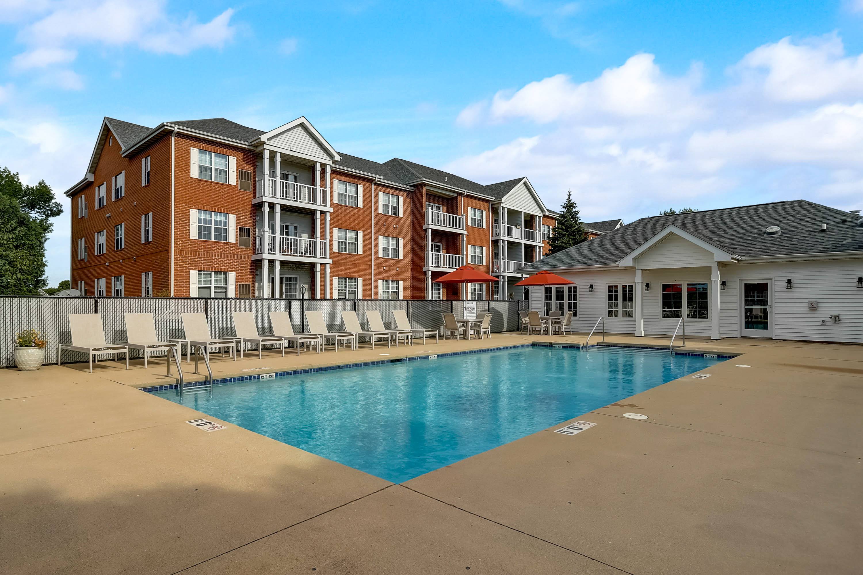A large swimming pool is surrounded by lounge chairs in front of a red brick apartment building.